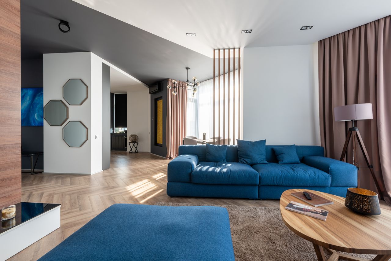 who-we-are Interior of spacious flat with blue sofa and pouf near wooden table placed on carpet near wall with stylish decorations